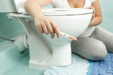 Closeup image of woman sitting on floor at bathroom and holding positive pregnancy test