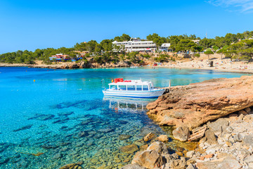 White and blue colour ferry boat for tourists mooring in Cala Portinatx bay, Ibiza island, Spain