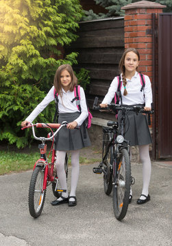Two smiling girls in school uniform walking with bicycles on street