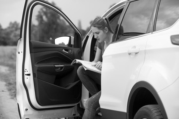 Black and white image of young woman sitting in car searching for road on map