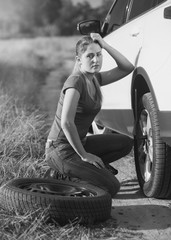 Black and white image of sad woman sitting at broken car and trying to change flat tire
