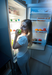 Young woman in pajamas eating pizza at open fridge at night