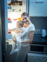 Smiling woman standing with her baby son on kitchen at night