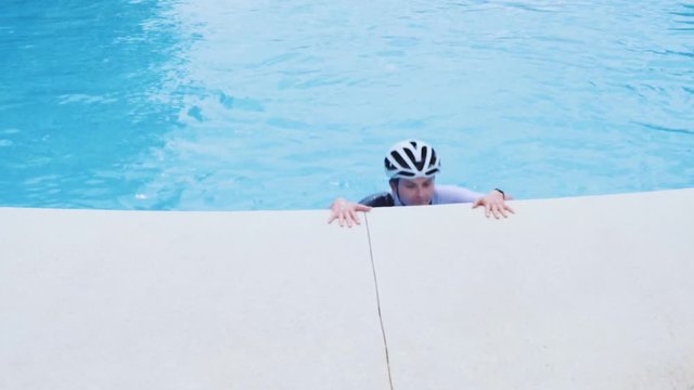 Tired happy cyclist woman in black and white bicycle clothes, helmet with blonde hair gets out from swimming pool after training and refreshment with cold fresh water. Girl breathes heavy and smiles.