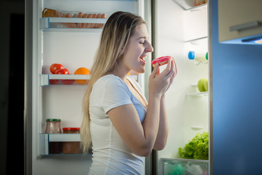 Portrait of young woman taking donut from refrigerator at night. Concept of unhealthy eating