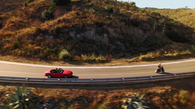 Epic aerial view from bird eyes on mountain serpentine road at sunny beautiful day, man in small red vintage convertible car chasing girl on retro motorbike at sunset summer day
