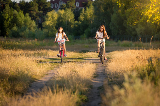 Two teenage girls riding bicycles on meadow at sunset