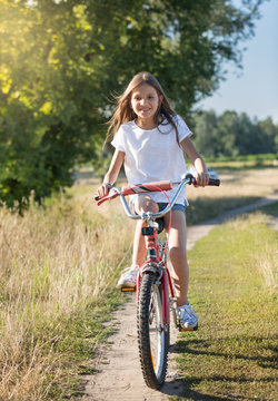 Cheerful girl with long hair riding her bicycle on dirt road at meadow
