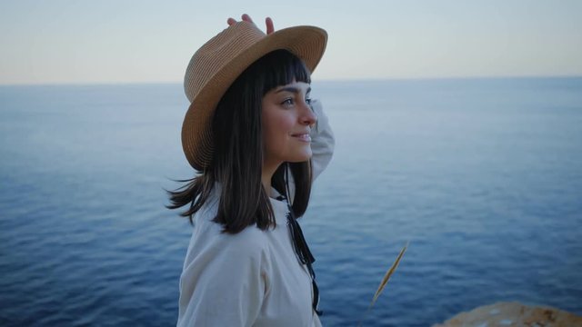 Portrait of beautiful smiling brunette girl wearing summer straw hat and white cotton dress, walking in rocks next to ocean backround at evening