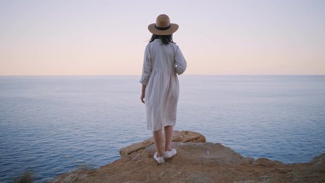 Rear view on shy and dreamy amish girl in simple white cotton dress and straw hat, standing on the edge of rock cliff and looking far away in ocean sea, waiting someone. Wind slightly swings her dress