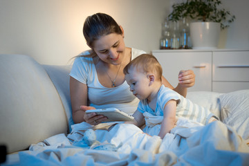 Baby boy sitting on bed with mother and using digital tablet at night