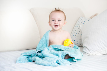 Freshly bathed baby boy covered in blue towel sitting on big sofa
