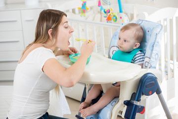 Smiling woman feeding her baby boy in highchair at living room