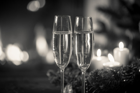 Closeup black and white image of two glasses of champagne in front of Christmas wreath and burning fireplace