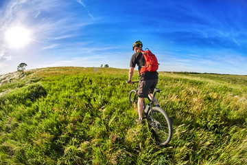 Fototapeta premium Young cyclist riding mountain bicycle through green meadow against beautiful sky.
