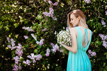 Obraz na płótnie Canvas Blonde girl with bouquet of wild flowers outdoors A model in a turquoise dress against a background of a lilac bush.