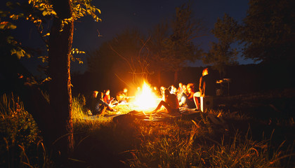 The company of young people are sitting around the bonfire and singing songs - obrazy, fototapety, plakaty