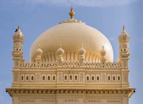 Mysore, India - October 26, 2013: Upper structure and dome of cream yellow Tipu Sultan mausoleum under blue sky. Golden Kalasam on top. One black bird.