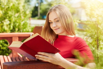 Outdoor portrait of young teenage girl with fair hair dressed in red blouse sitting at wooden bench and reading her favourite book admiring summer weather and sunshine. Relaxation and rest concept