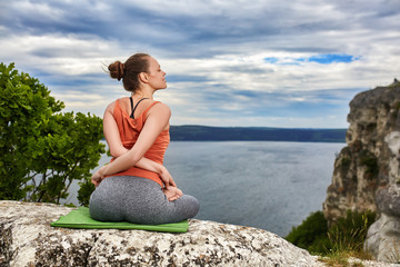 Naklejka na ściany i meble Rear view of young woman in yoga pose sitting on the rock above river.