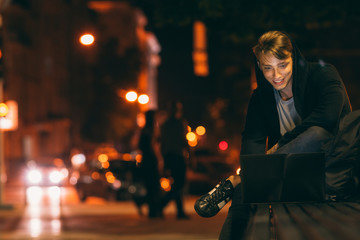 Young man with laptop in night city, free space. Student working on his computer in park in night time. Smiling guy searching information at place with free wi-fi