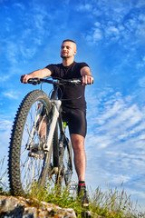 Naklejka premium Bottom view of young cyclist standing with bike on rocks against blue sky.