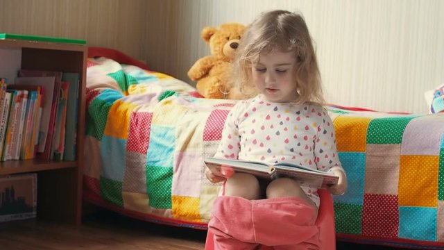 Little girl sitting on the potty beside the bed and reading a book.