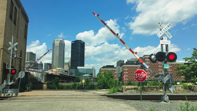Bicyclists in helmets wait for the train crossing barriers to raise before crossing the tracks. The Pittsburgh skyline is in the distance.  	