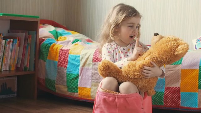 Little girl sitting on the potty beside the bed and plays with a Teddy bear.