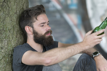 Handsome hipster guy leaning against a tree drinking a beer in park