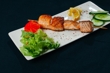 Shashlik of salmon with vegetables on white plate and black background