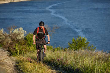 Portrait of the young cyclist riding mountain bike uphill above beautiful blue river.