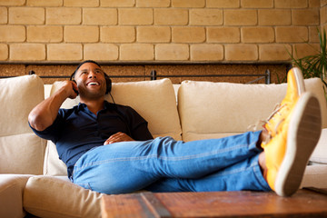 Cheerful young black man lying on sofa with headphones and listening to music
