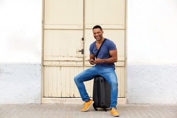 Full length happy african man sitting on a suitcase with mobile phone and smiling