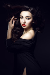 Portrait of glam dark-haired model with perfect make up and flying wavy silky hair wearing golden luxurious necklace and black side split slit top. Isolated on black background. Studio shot