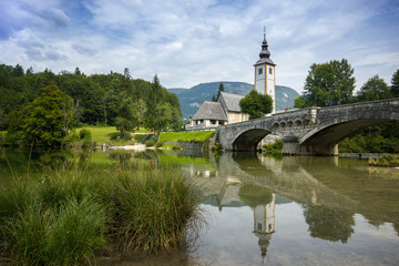 Fototapeta na wymiar Old church and bridge mirroring in river, Bohinj lake, Slovenia