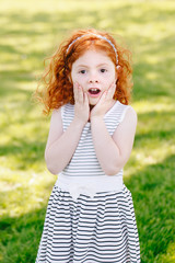 Portrait of cute adorable surprised  little red-haired Caucasian girl child in striped dress in park outside, playing  crying screaming in fear, happy lifestyle childhood concept