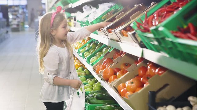 Cute baby girl is picking up tomato pack from a shelf in vegetables department in a supermarket