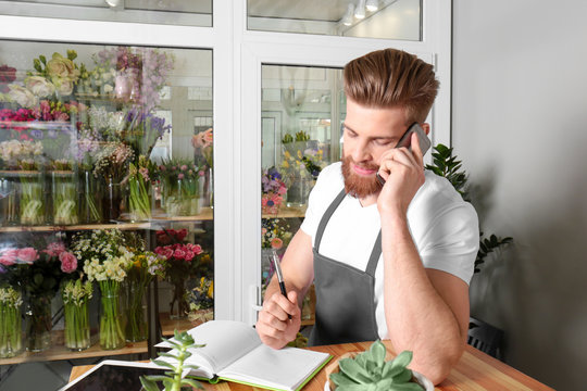 Young handsome florist writing order while talking by mobile phone in flower shop