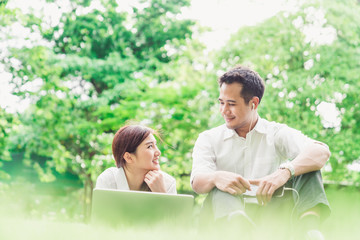 Young Asian lovely couple or college students look at each other in the garden or park, using laptop computer and smartphone, listening to music. Love, relationship, relaxing casual lifestyle concept