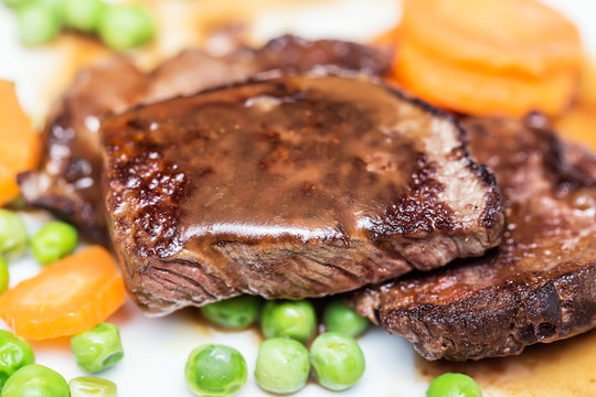 Juicy beef meat steaks with sauce, potatoes, carrots and peas on white plate, close-up. Selective focus