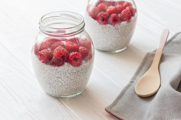 Glass jars of pudding from chia seeds with coconut milk and raspberry berries in white wooden table.  Linen towel and spoon.