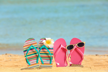 Naklejka premium Colorful flip-flops, sunglasses and flower on sand at sea shore. Vacation concept
