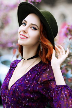 Outdoor close up portrait of young beautiful happy smiling woman looking at camera, posing in street. Model wearing stylish green fedora hat, purple dress, choker necklace. City lifestyle
