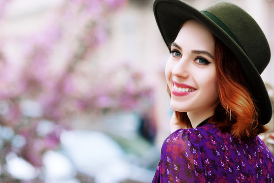Outdoor close up portrait of young beautiful happy smiling woman looking aside, posing in street. Model wearing stylish green hat, purple dress. Female fashion concept. Copy, empty space for text