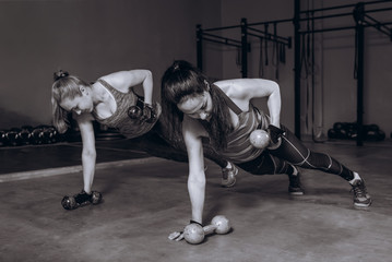 Two fit women in gym doing fitness exercises with dumbbells staying in plank pose, black and white