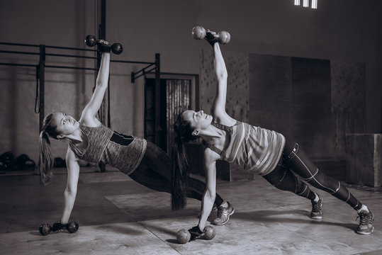 Two fit women in gym doing fitness exercises with dumbbells staying in plank pose, black and white
