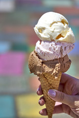 Chocolate, vanilla and berry ice cream scoops in waffle cone held by female hand against colorful background