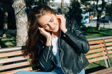 The beautiful girl listens to music with white earphones and enjoys. She sits on a bench in the solar park. On the girl a black leather jacket, light trousers and an undershirt.