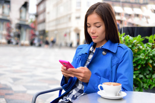 Cafe woman texting sms on phone app drinking cappuccino coffee at outdoor street terrasse in european city. Europe travel lifestyle. Asian businesswoman in blue trench coat.
